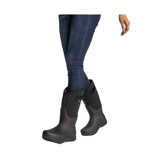 Black knee-high rain boots with pull-on cuffs and thick tread soles, shown on a model with jeans