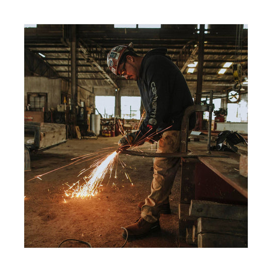 Black hooded sweatshirt with Carhartt logo on sleeve, worn by a worker grinding metal in a workshop