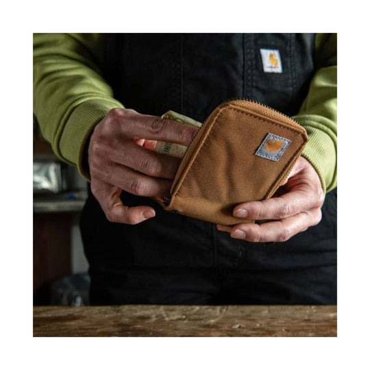 Brown zippered canvas wallet being held open, with a small square logo patch on the front.