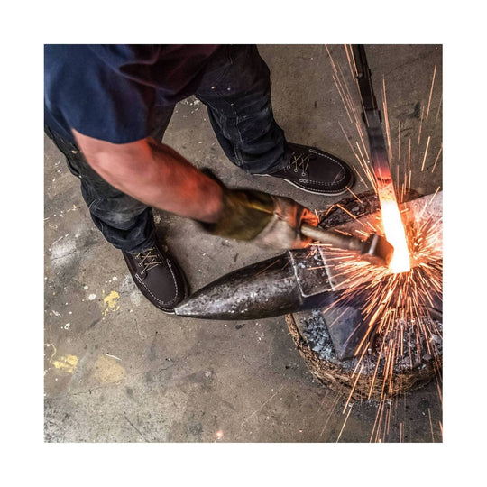 Brown leather moc toe work boots with white stitching and laces, worn while blacksmithing beside sparks