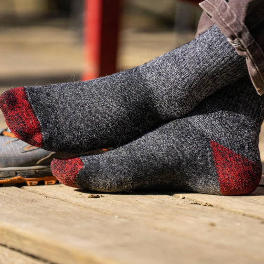 Gray wool hiking socks with red toes and heels worn over a shoe on a wooden surface.