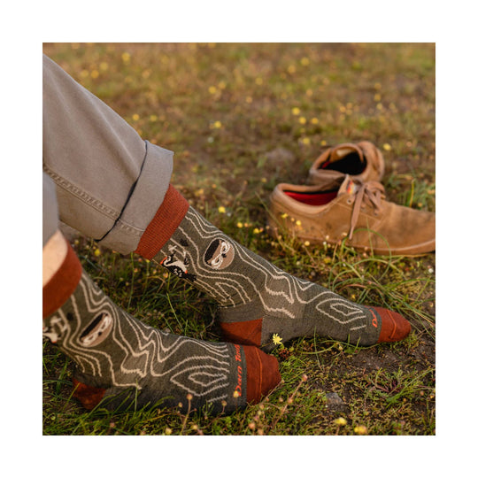 Patterned crew socks in olive and rust, worn with rolled khaki pants, shown beside a pair of tan shoes on grass.
