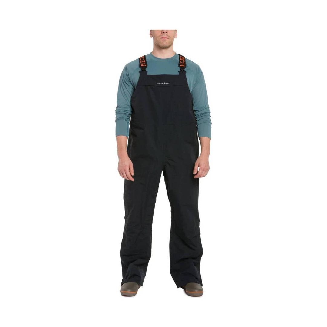 Black bib overalls with shoulder straps worn over a teal long-sleeve shirt