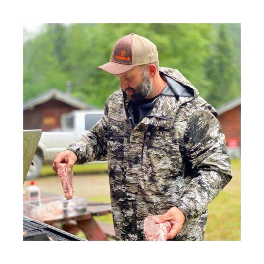 Camouflage hooded jacket with brown and tan baseball cap, worn while grilling outdoors