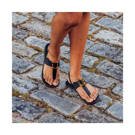 Black thong sandals with tortoiseshell buckles worn on a cobblestone path