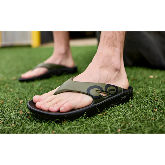 Olive slide sandal with black sole and large side logo, worn on grass