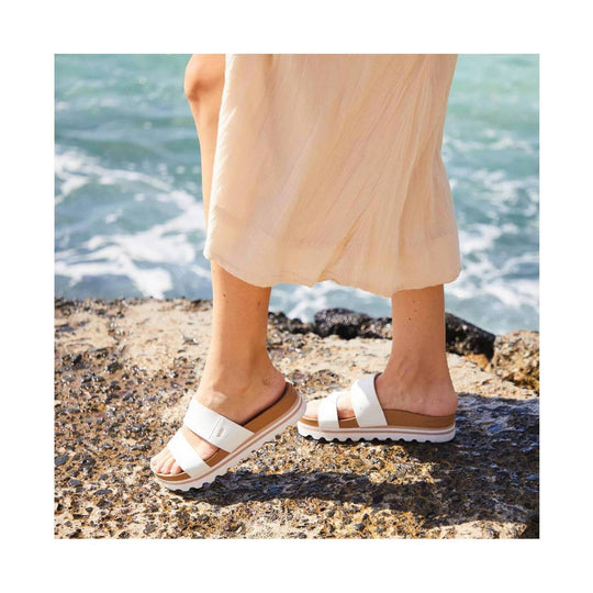 White and tan platform slide sandals with wide straps, worn outdoors on rocks by the sea.