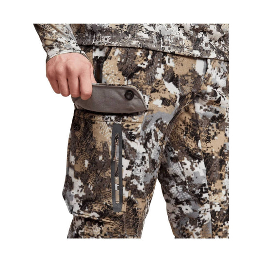 Camouflage outdoor pants with a zip cargo pocket and a hand pulling out the flap