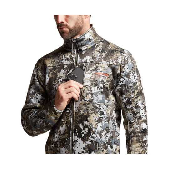 Men's camo zip-up jacket with a high collar and chest pocket, shown on a model holding a phone