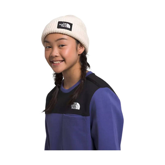 Cream knit beanie with a black The North Face patch, shown on a child wearing a purple-and-black top.