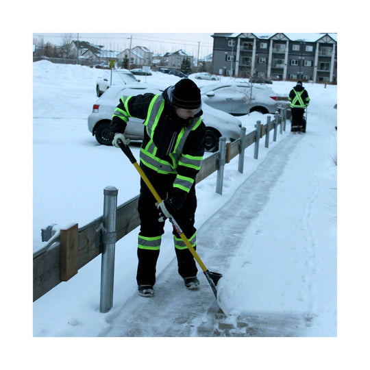 Two workers in reflective safety jackets shoveling snow from a sidewalk in a parking lot