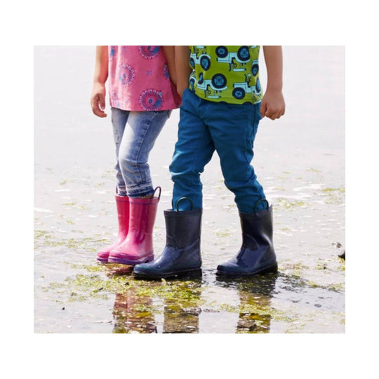 Two children wearing tall rubber rain boots standing in shallow water, one pink and one navy.
