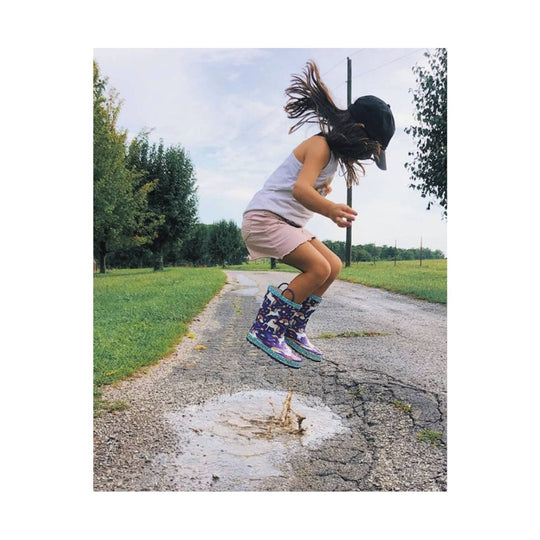 Purple unicorn-print rain boots worn by a child jumping in a puddle